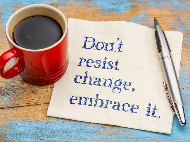 A cup, napkin with "don't resist change, embrace it," written on it, and a pen on top a wood surface.