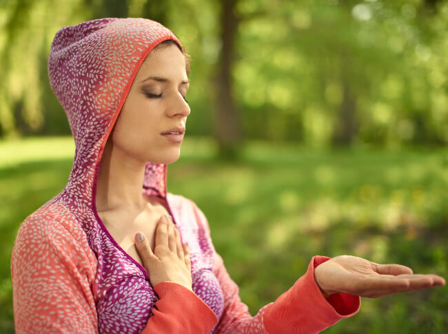 a woman outside with her eyes closed, one hand on her heart and the other out with the palm up.