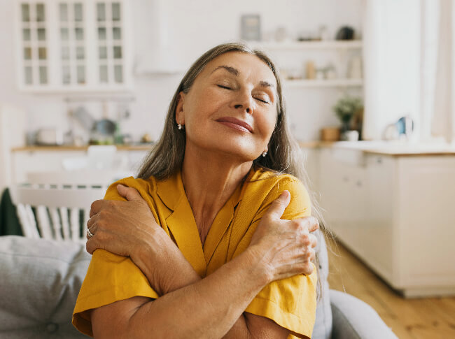 An older woman sitting on a couch in a home hugging herself.