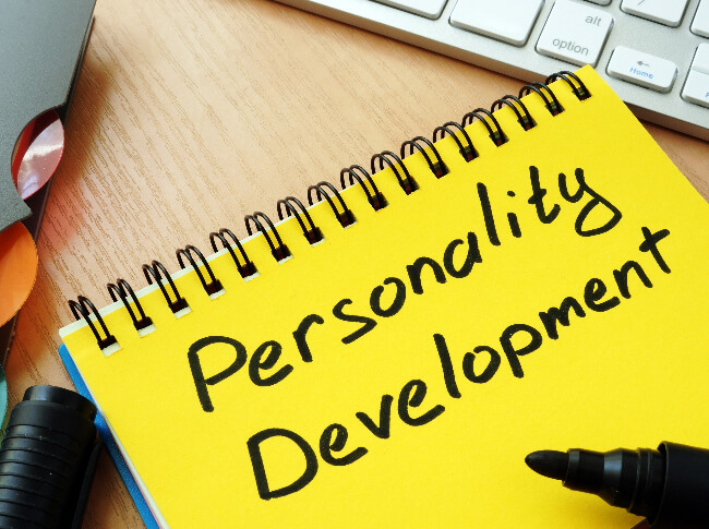 image with a yellow pad that says personality deveopment written with a marker (which is on top the pad) and there is a part of  keybard in the shot as well as a folder and the marker's cap.