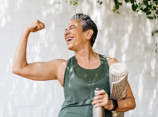 A woman with a yoga matt rolled under her arm and a water bottle in the hand - she's smiling beautifuly while making a muscle in celebration with her free  arm.
