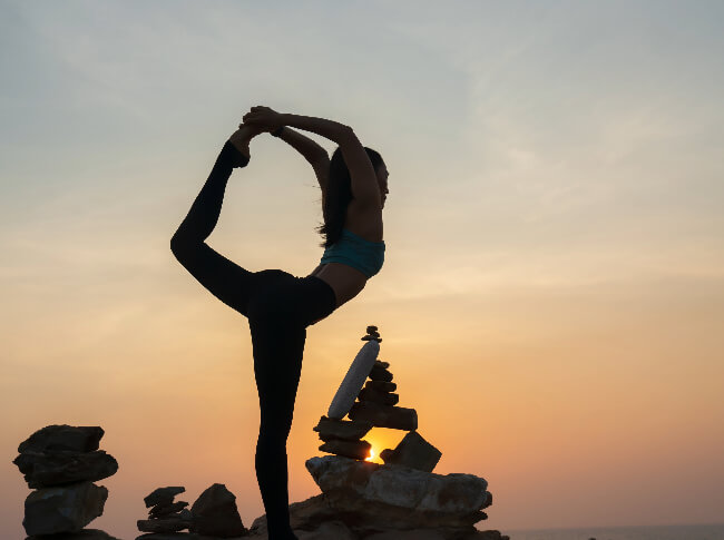 The silhouette of a woman doing joyga outside with stones around her and a beautiul sky background with the sunsetting or rising.