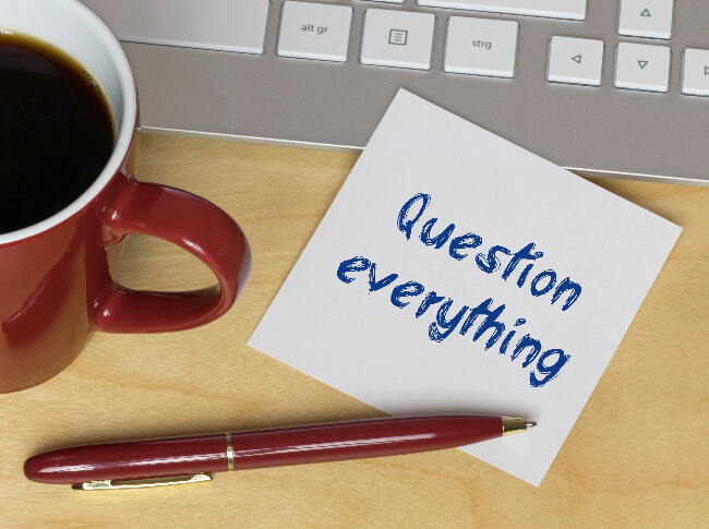 Cup, part of laptop,and coffee cup and pen ontop of a post it note that says, Question Everything.