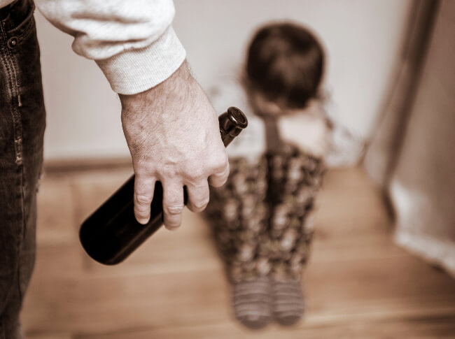 A little boy in the corner with his head down and a man with  bottle of alcohl in front of him