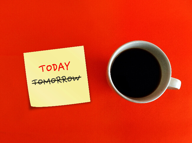 Photo with a sticky with a strikethrough on the word  TOMORROW  and with TODAY above it to replace TOMORROW. next to a cup of  coffee.