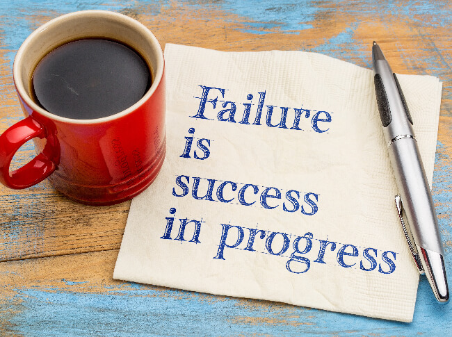 A wooden desk with a cup of coffee, a napkin that reads, "Failure is Success in progress," and  pen on topo of it.
